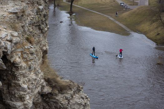 Two adults paddleboarding near cliffside on a river, aerial perspective.