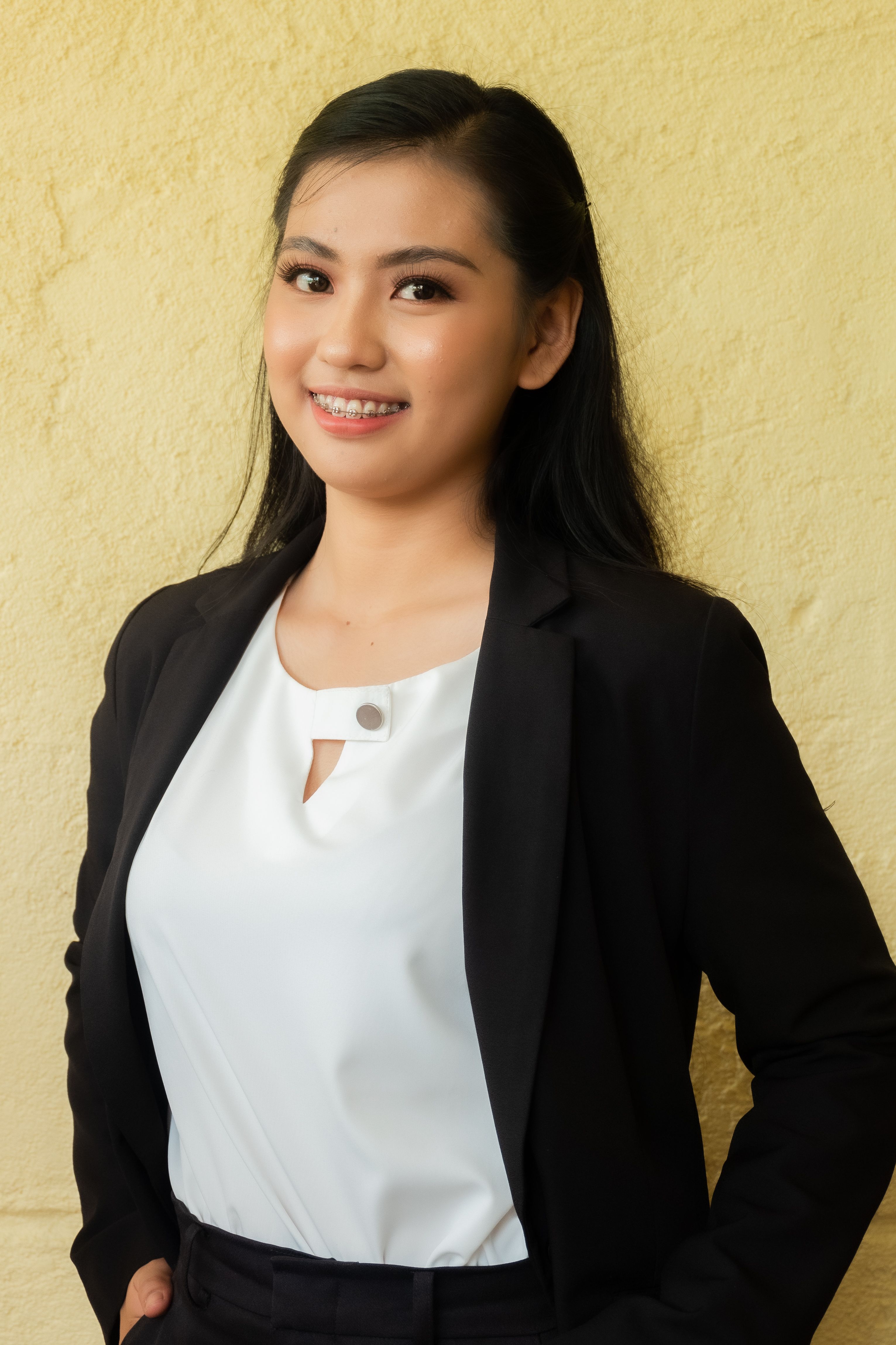 Confident Asian woman in a black blazer smiling against a beige background.