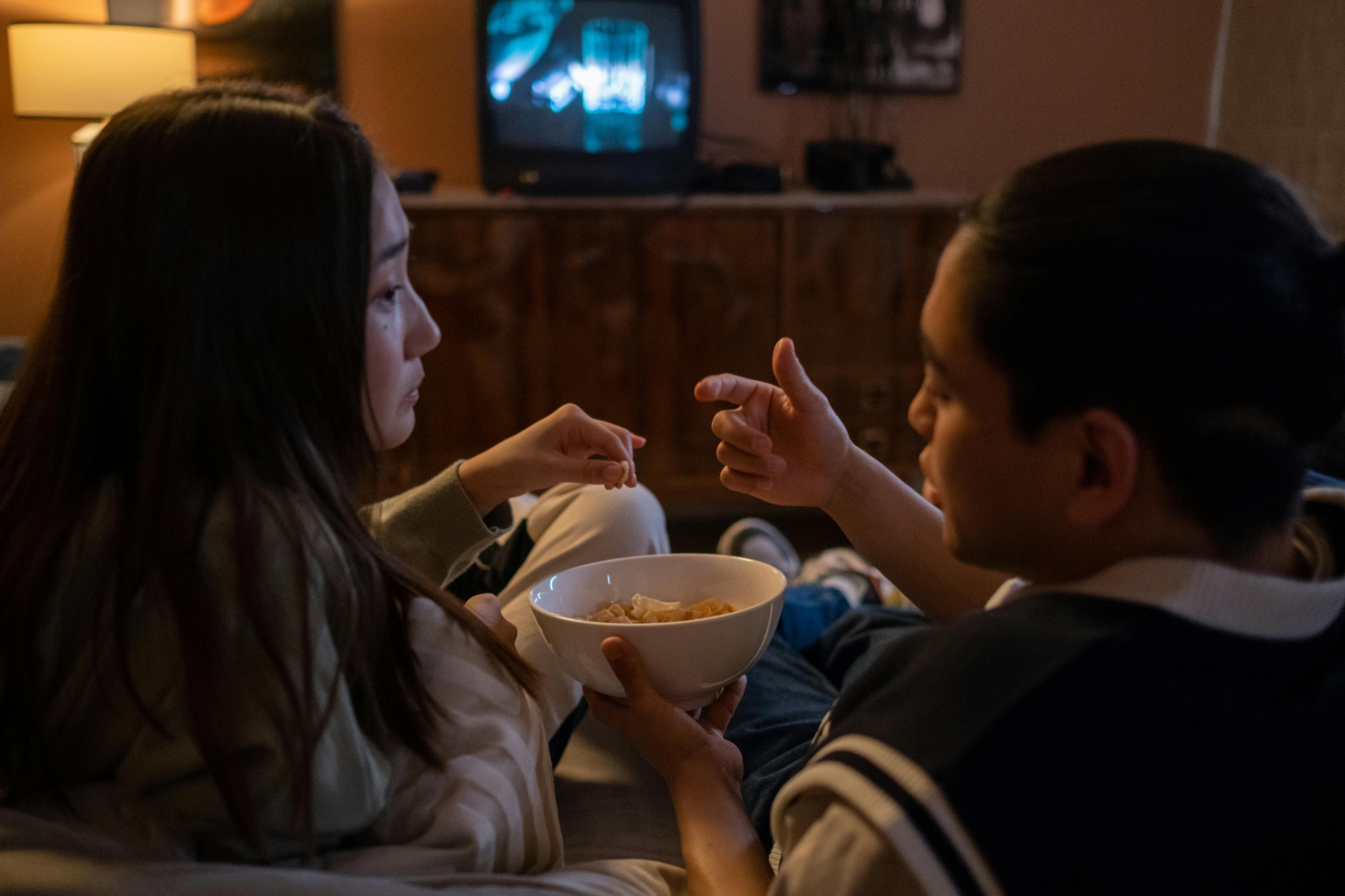 Asian couple enjoying snacks while watching TV at home during evening.