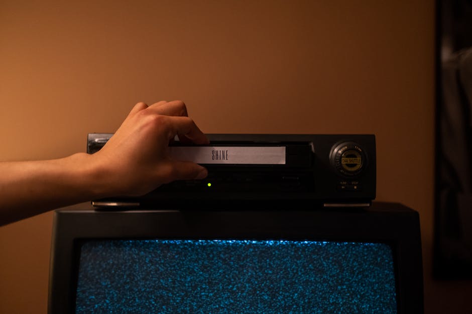 A hand inserting a VHS tape labeled 'SHINE' into a vintage player. Nostalgic home movie setup.