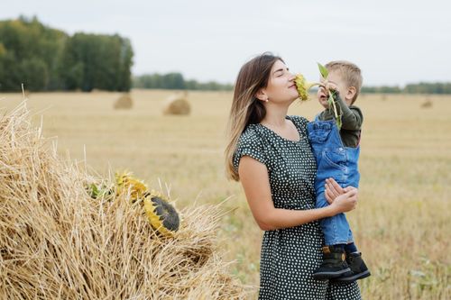 Free Mother holding child smelling a sunflower in a sunny field with hay bales. Stock Photo
