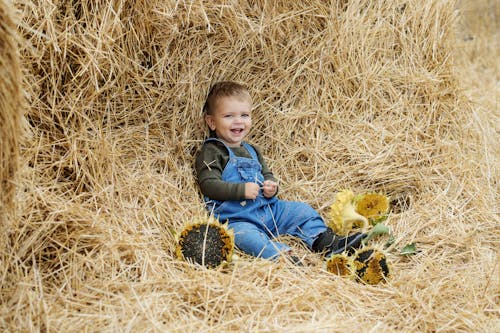 A Boy Wearing Long Sleeves and Denim Jumper Sitting on a Haystack