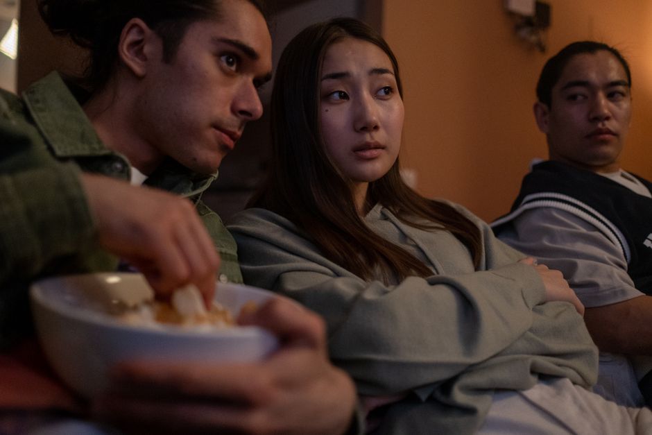 Three teenagers relaxing indoors watching TV, sharing snacks during a cozy movie night.