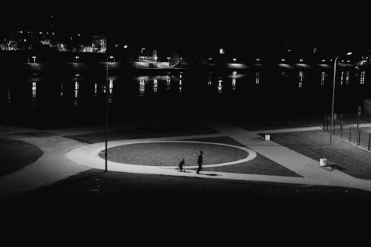 Black and white park at night with silhouetted figures walking, distant river reflections.