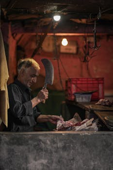 A butcher in a dimly lit shop preparing raw meat with a cleaver.