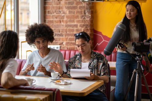 Free A diverse group of filmmakers working indoors in a cafe setting, capturing a scene with a camera and microphone. Stock Photo