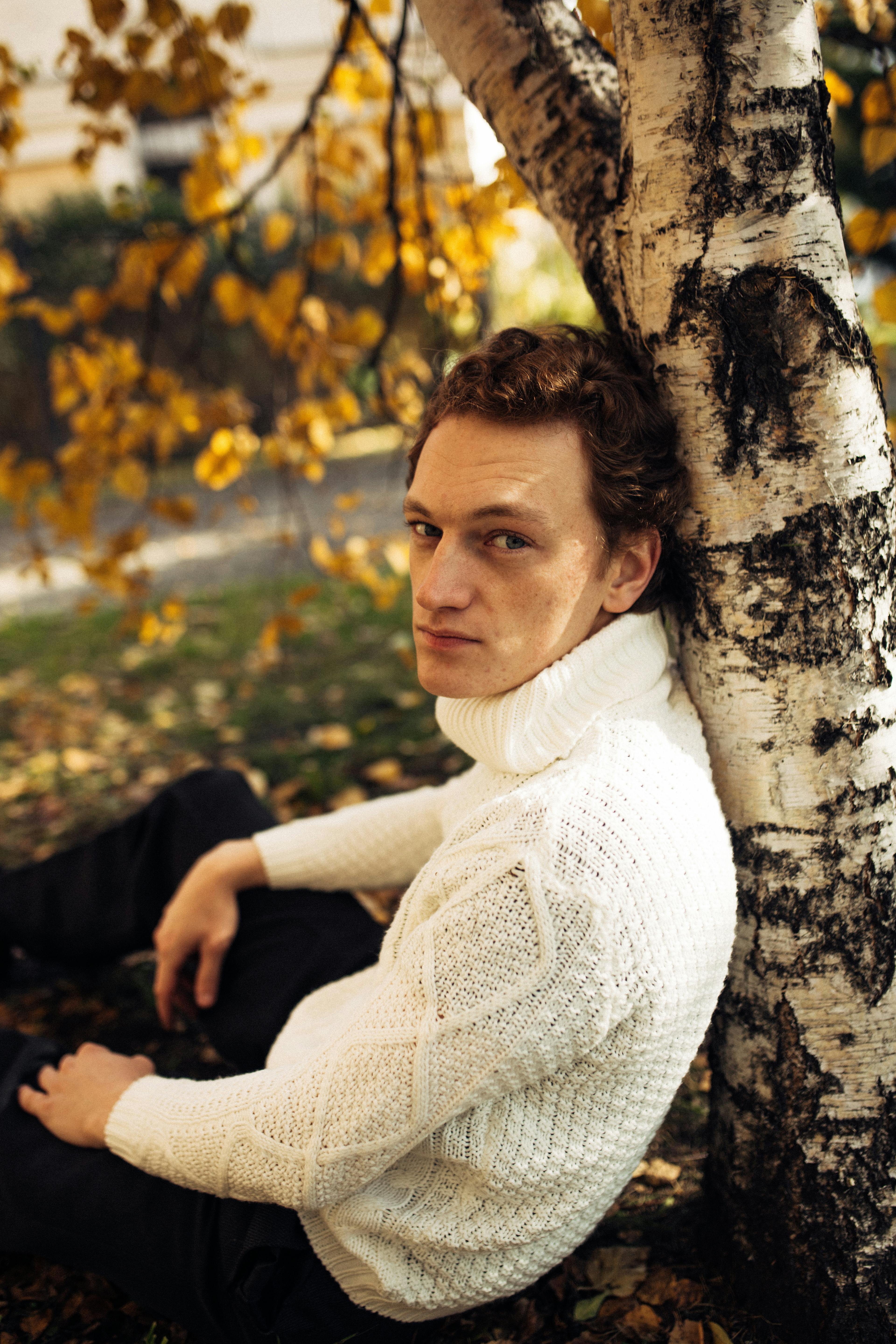 Man in a white sweater sitting under birch in autumn setting.