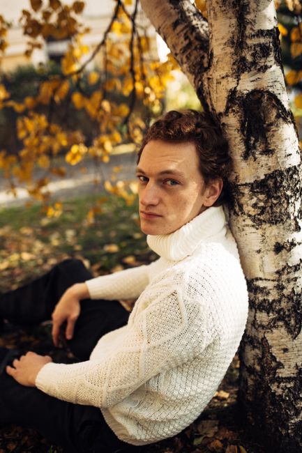 Man in a white sweater sitting under birch in autumn setting.