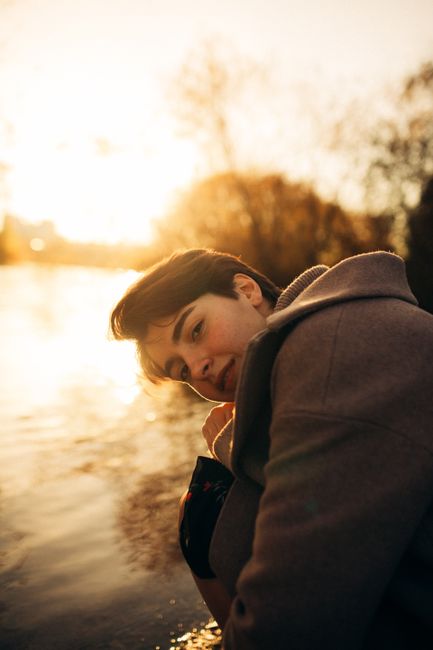 A young man in a coat kneels by a tranquil lake during sunset, exuding a serene and reflective mood.