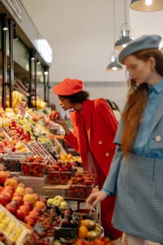 Stylish women exploring a vibrant indoor fruit market in chic outfits, offering a blend of fashion and freshness.