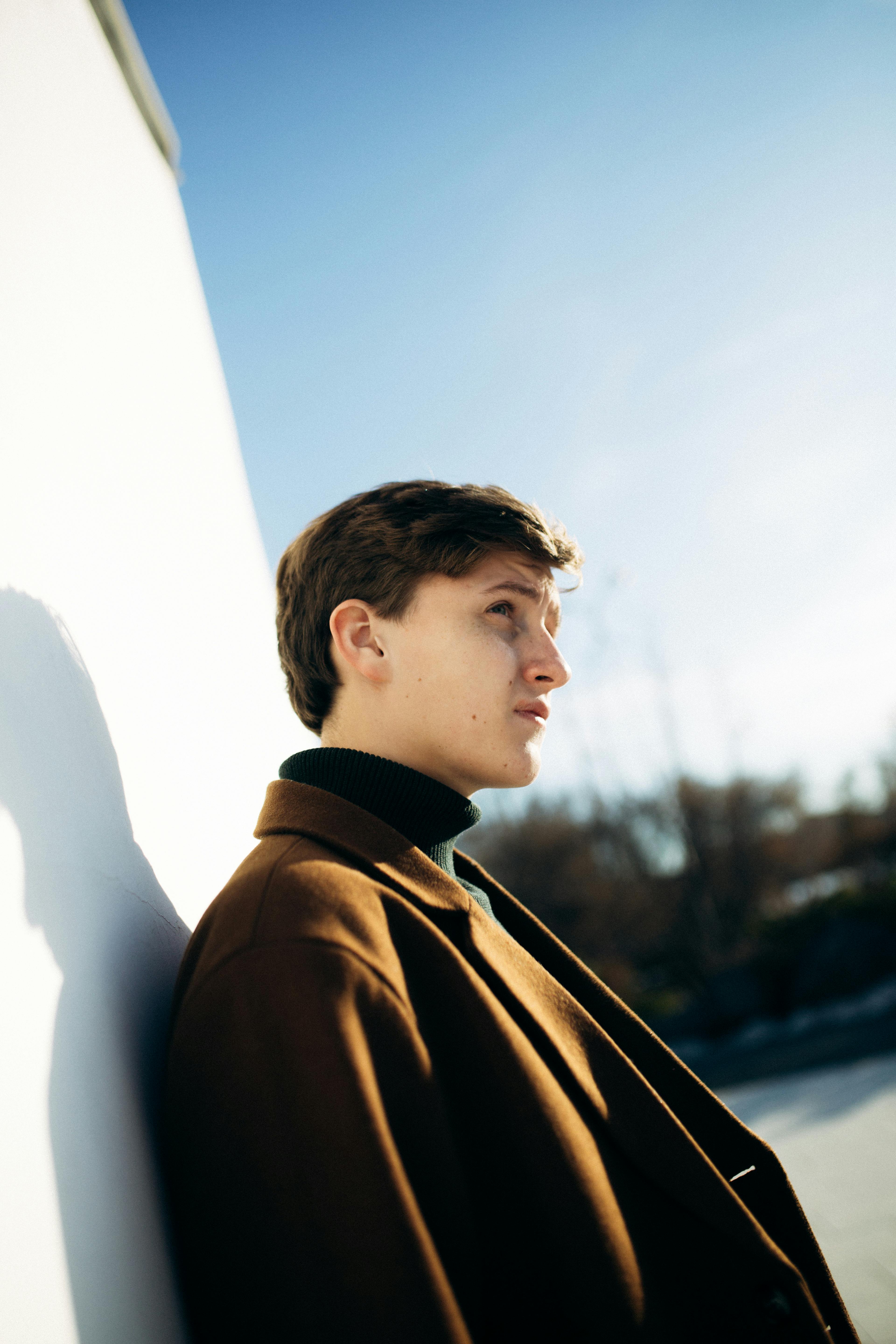 Portrait of a young man in a brown jacket leaning against a white wall under a clear sky.