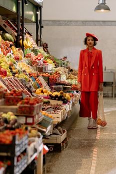 Fashionable woman in red outfit shopping for fruits at an indoor market, holding a mesh bag.