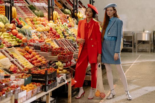 Stylish women in vibrant outfits shopping at a colorful fruit market.