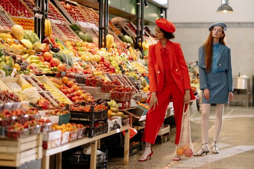 Two stylish women in fashionable attire shopping at a colorful fruit market, capturing urban lifestyle.