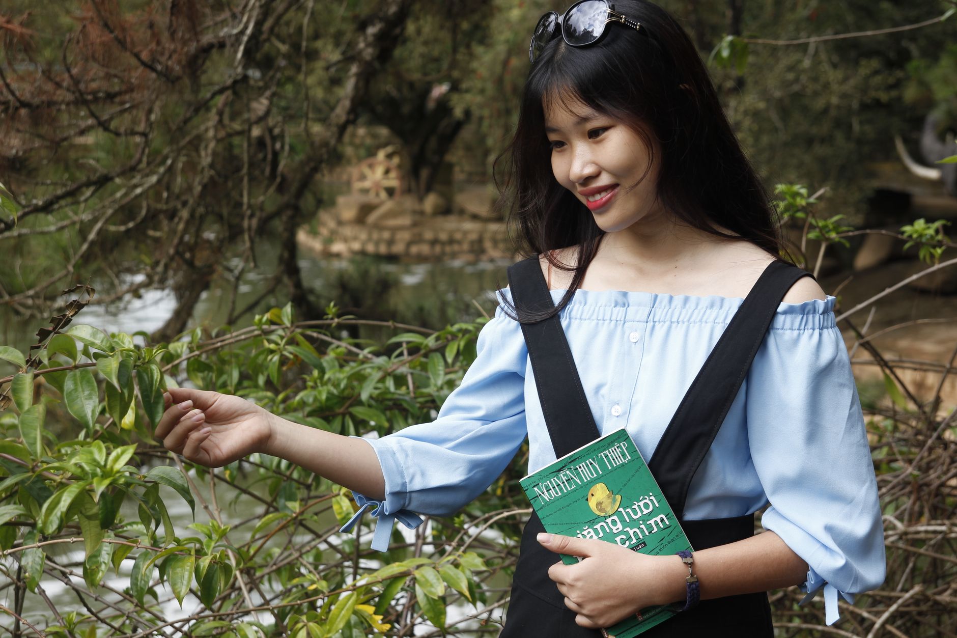 Smiling woman in a park holding a book, enjoying a sunny day outdoors.