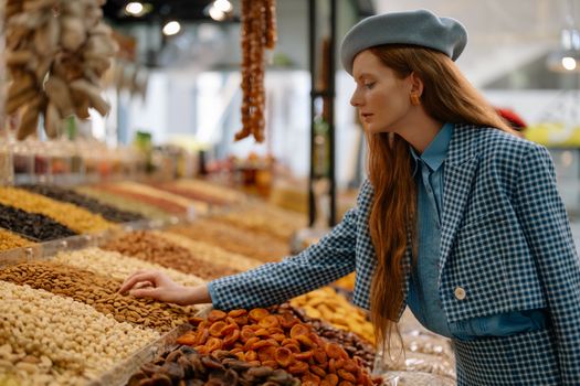 Woman in stylish outfit selecting dried fruits and nuts at a market display.
