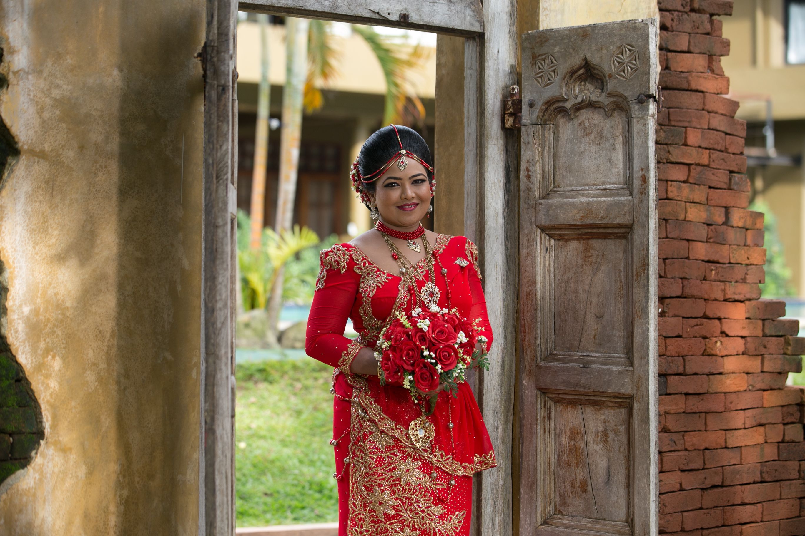 Bride in vivid red saree framed by a carved wooden doorway