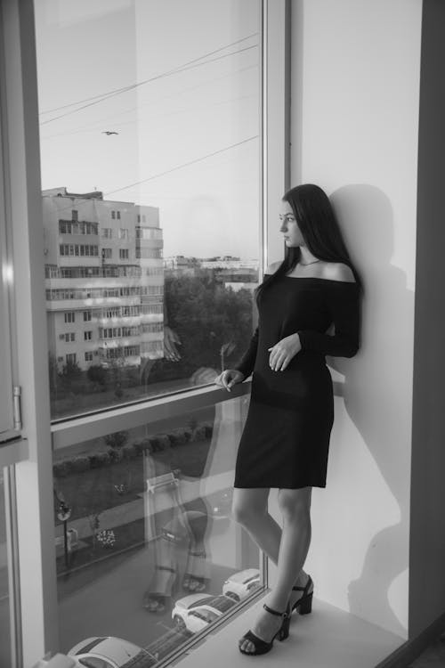 Monochrome Photo of a Woman in a Dress Standing Beside a Window