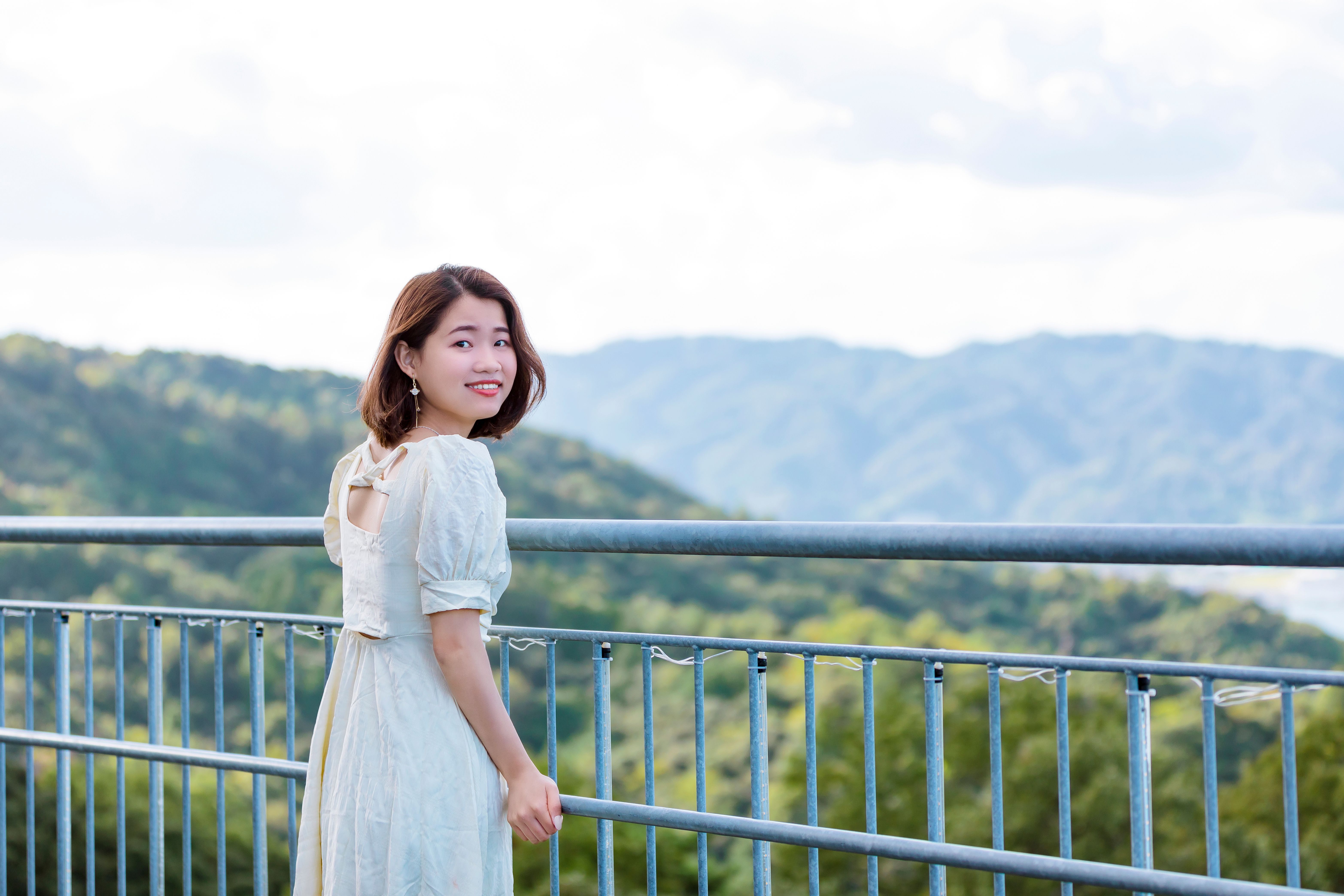 A smiling woman in a white dress on an outdoor balcony with scenic mountain views.