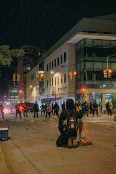 A protester kneels on a city street at night facing a line of police under streetlights.