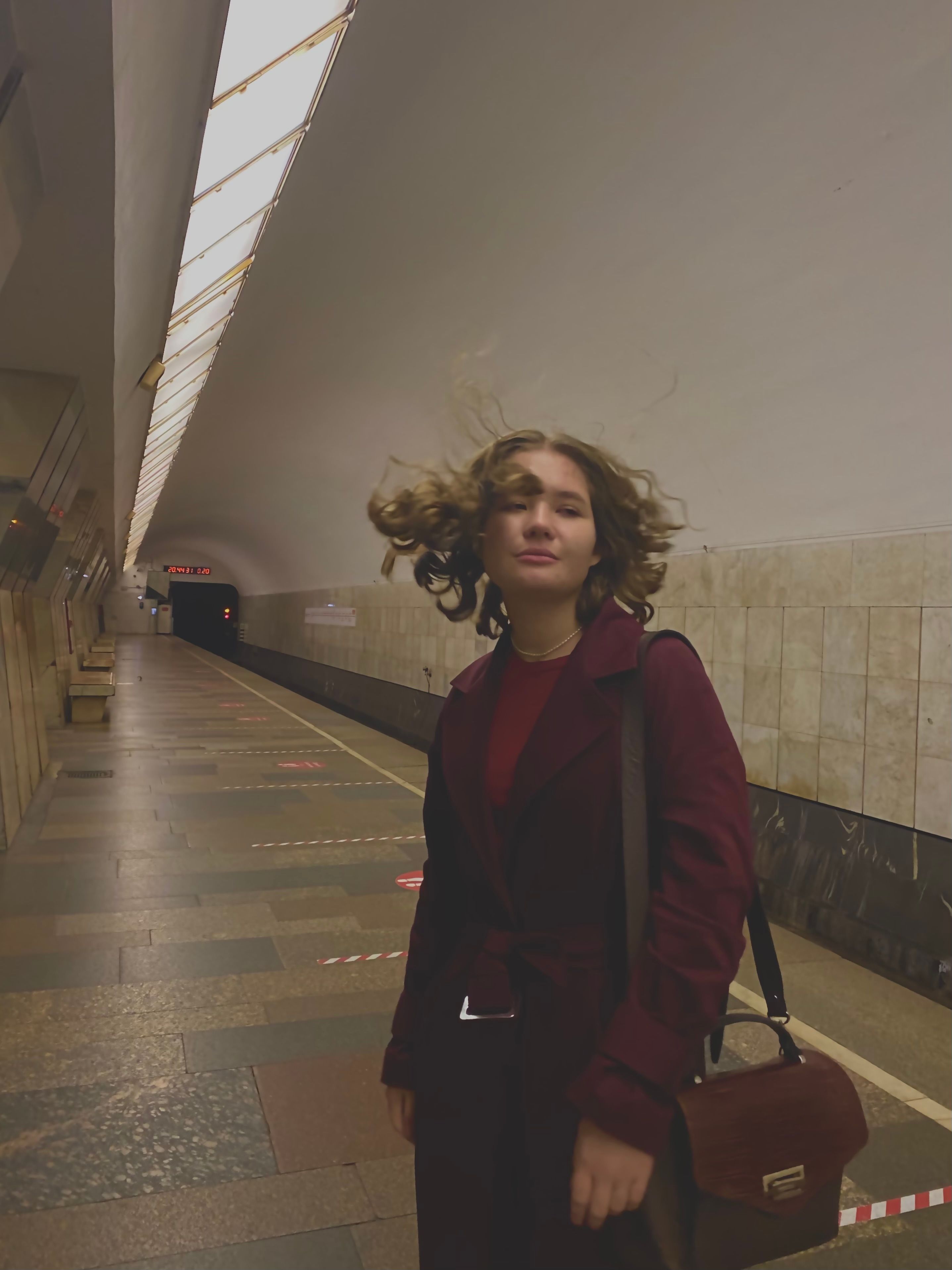 Portrait of a woman in a maroon blazer with a sling bag in an underground subway corridor.