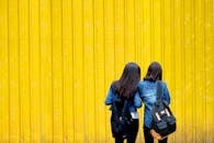 两名背着背包、穿着牛仔夹克的女性站在一面鲜艳的黄色墙壁前