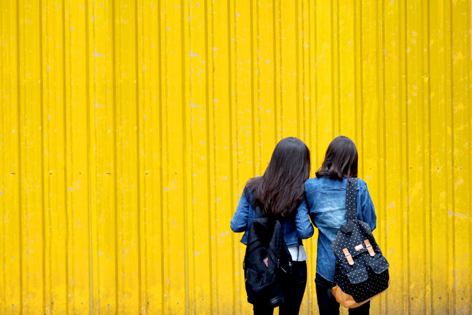 Two women wearing denim jackets and backpacks stand against a vibrant yellow wall.