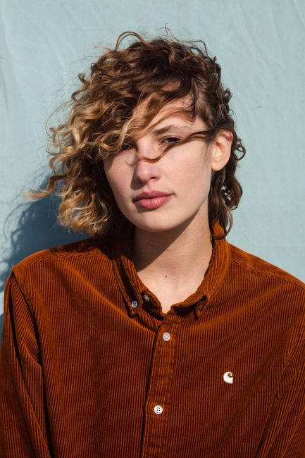 Close-up portrait of a woman with curly hair in a brown corduroy shirt against a blue background.