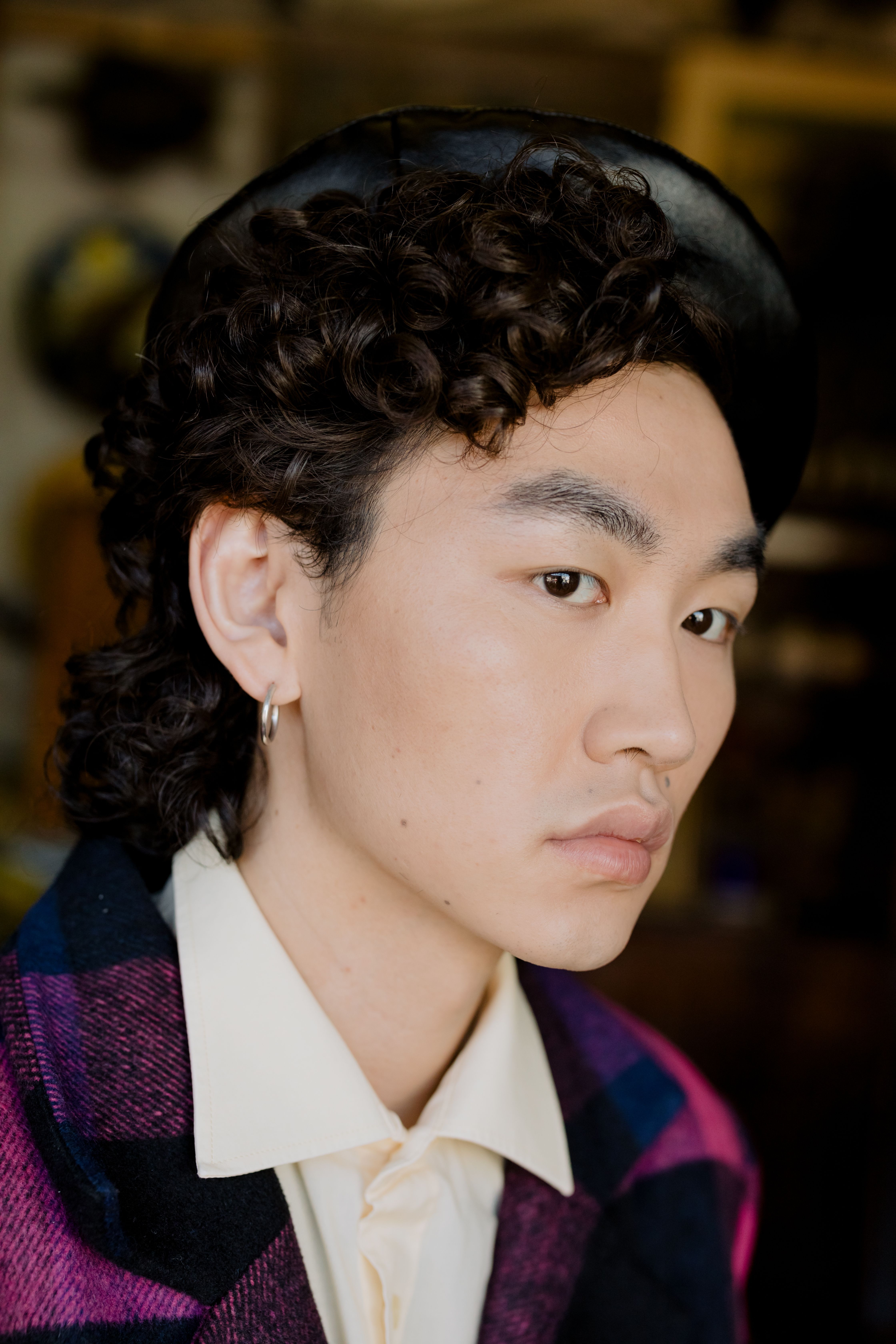 Close-up portrait of a stylish Asian man with curly hair wearing a fashionable jacket and hat.
