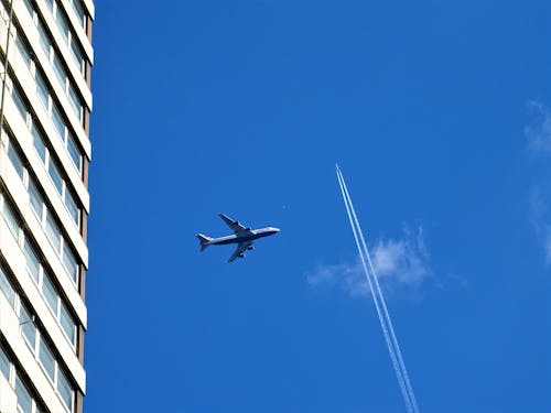 Low Angle Fotografie Van Een Wit Vliegtuig In De Lucht In De Buurt Van Een Wit Betonnen Gebouw En Een Wit Contrail