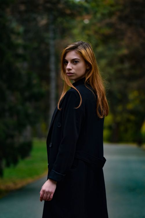 Free A woman in a black coat stands outdoors with blurred foliage in the background, creating a serene atmosphere. Stock Photo
