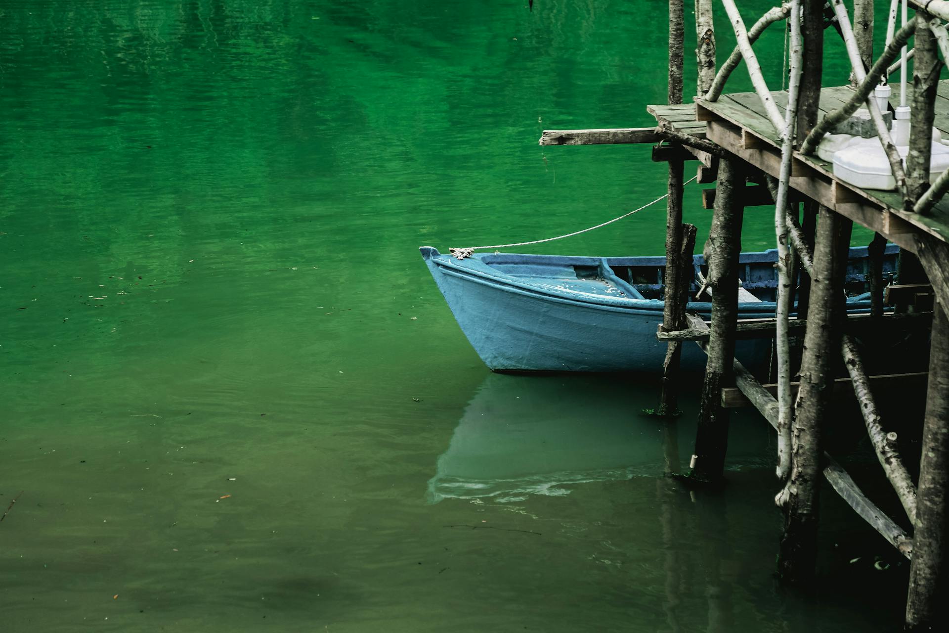 A tranquil scene of a blue boat tied to a rustic wooden dock over calm green waters.
