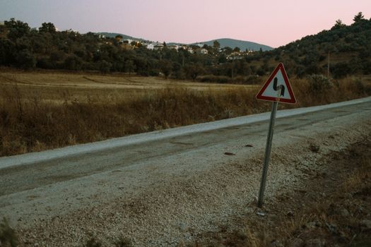 A rustic rural road with a double curve road sign and scenic landscape at dusk.