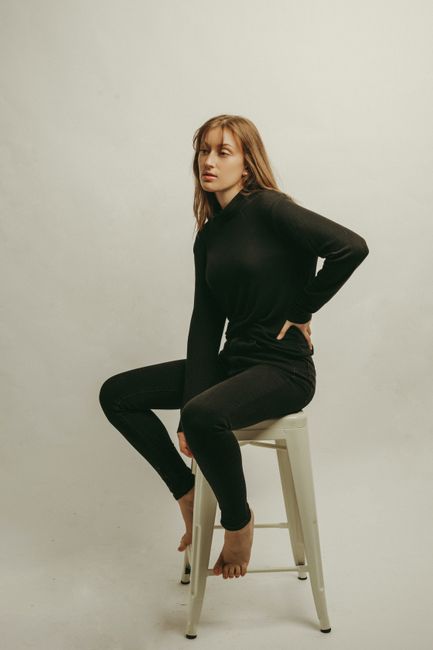 A woman in black attire sits on a stool in a minimalist studio, exuding serenity.