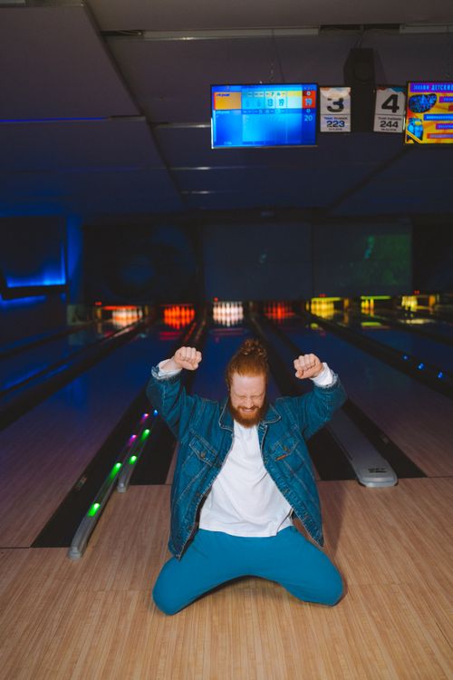Free A man kneeling with fists raised in celebration at a dimly lit bowling alley. Stock Photo