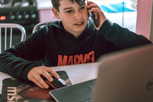 A Boy in Black Hoodie Using a Phone and a Laptop
