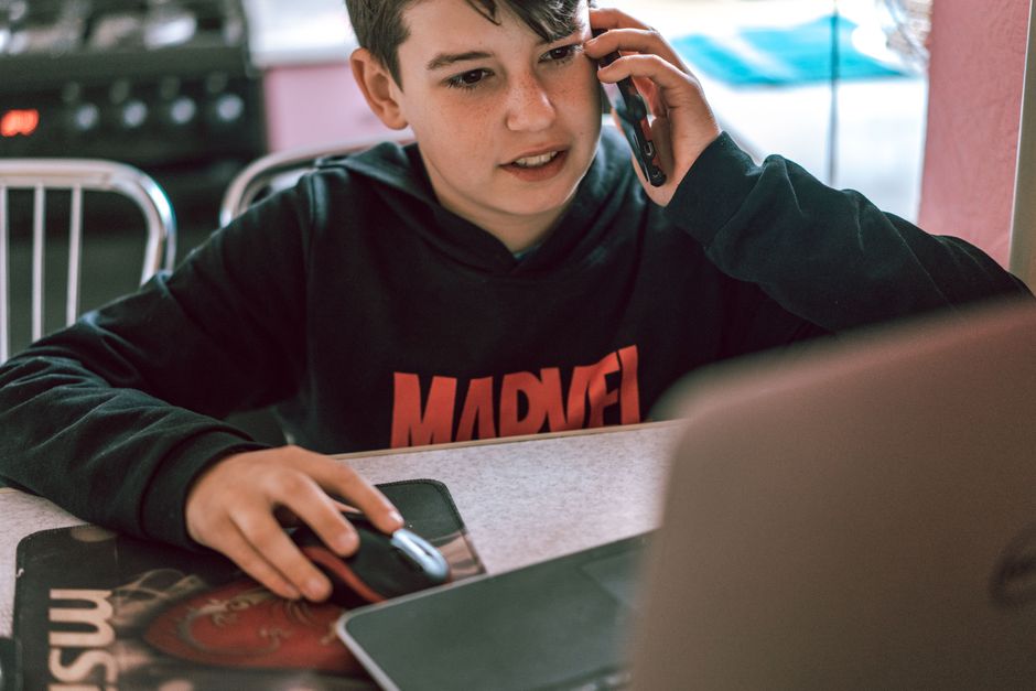 Teenage boy using a phone and laptop, engaging in multitasking indoors.
