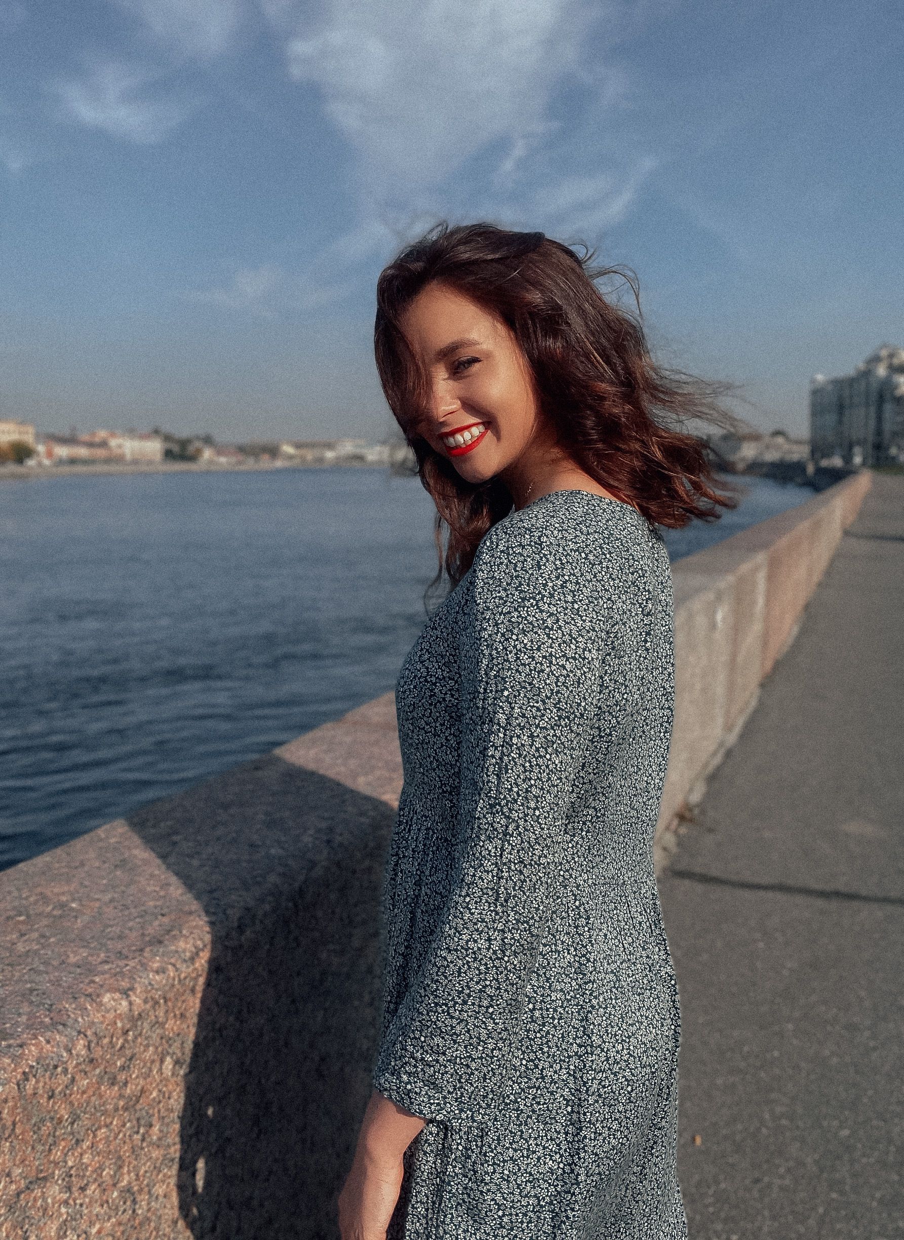 A woman in a patterned dress smiling by a city waterfront under a sunny sky.