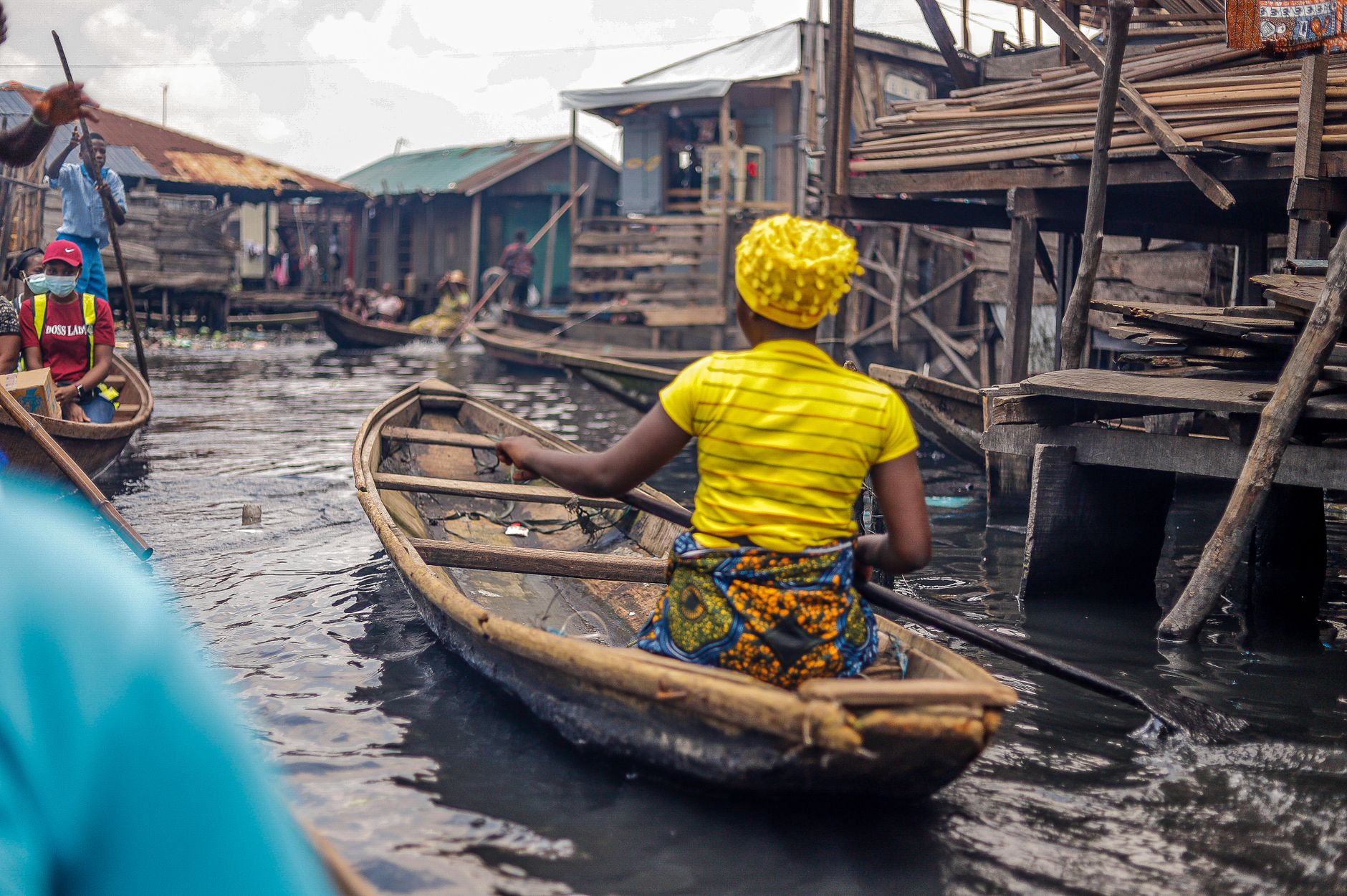 An African woman paddles a canoe through a bustling urban waterway in a traditional setting.