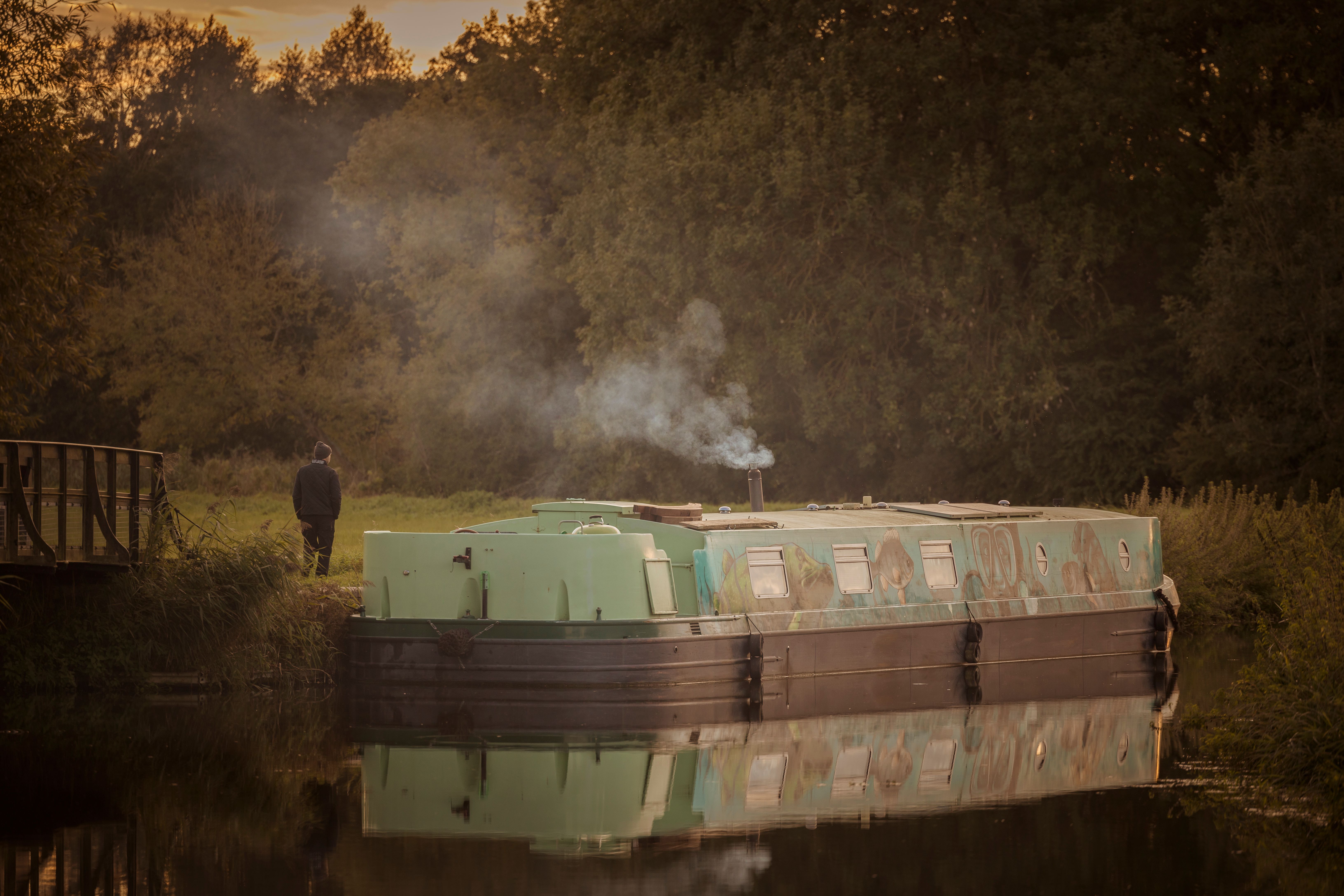 Serene image of a barge moored by a riverbank at sunset with smoke rising into the forested background.