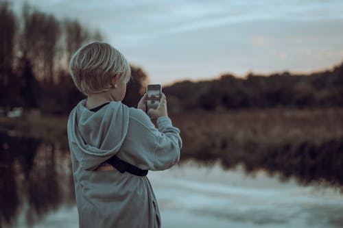Boy in Gray Hoodie Taking Picture of the Lake on Mobile Phone During Sunset 