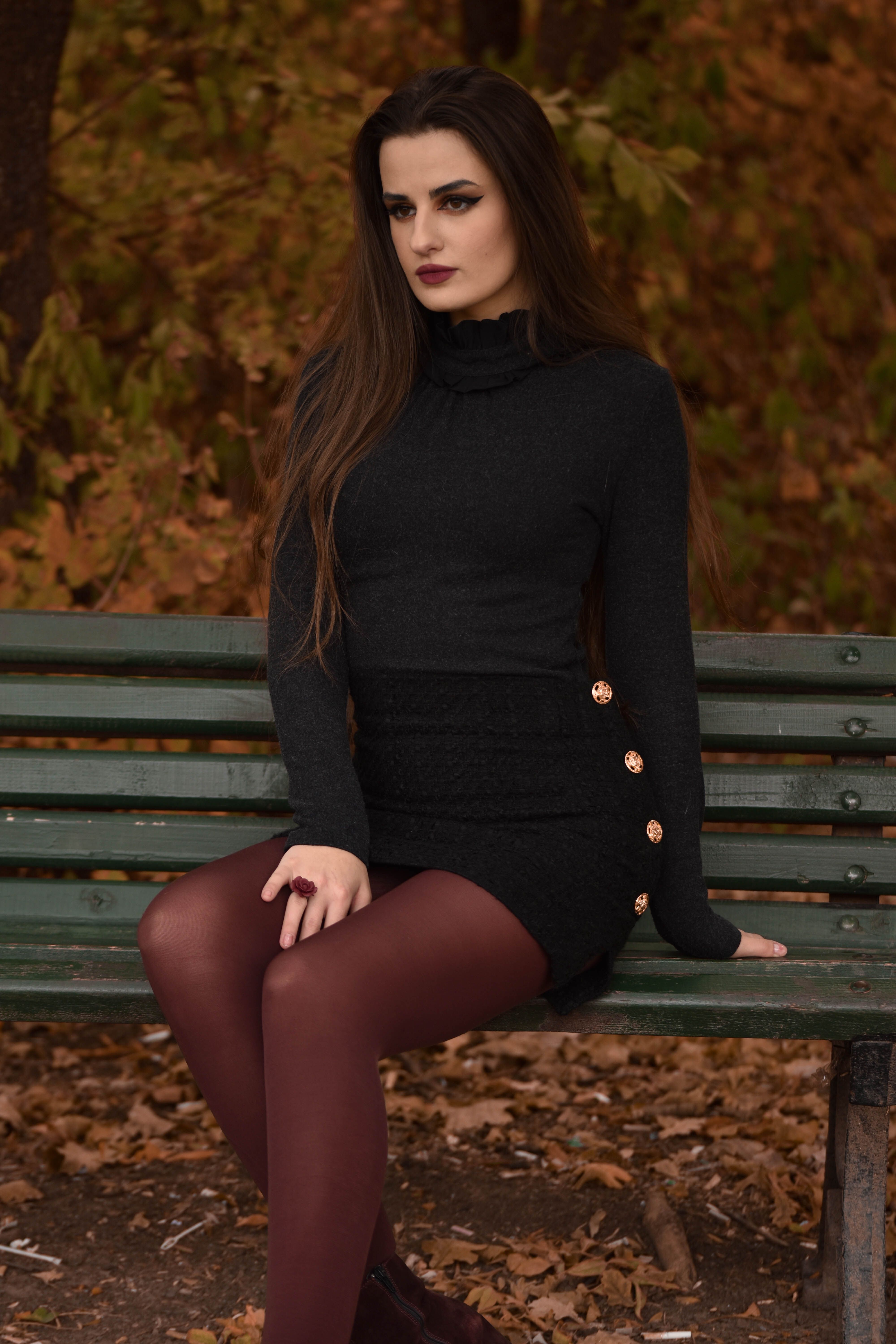 A fashionable woman sitting on a green bench in a park during fall, surrounded by vivid autumn foliage.