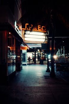 A cinematic urban street scene at night featuring a neon-lit theatre marquee and vibrant city lights.