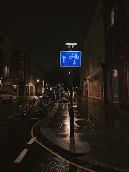 Rain-soaked London street at night featuring a bicycle lane sign and wet sidewalks.
