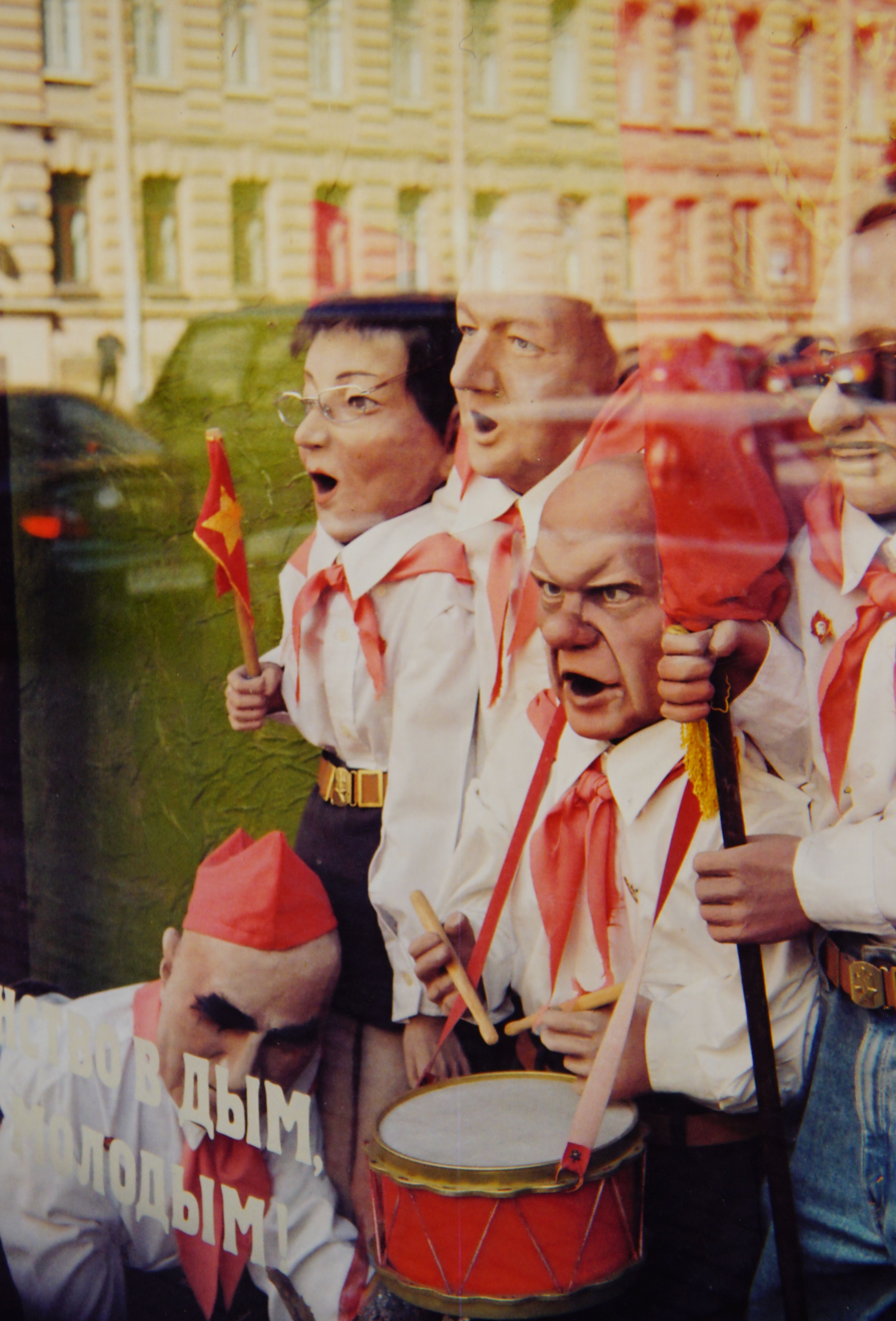 Surreal depiction of masked figures in a reflective window holding drumsticks and flags.