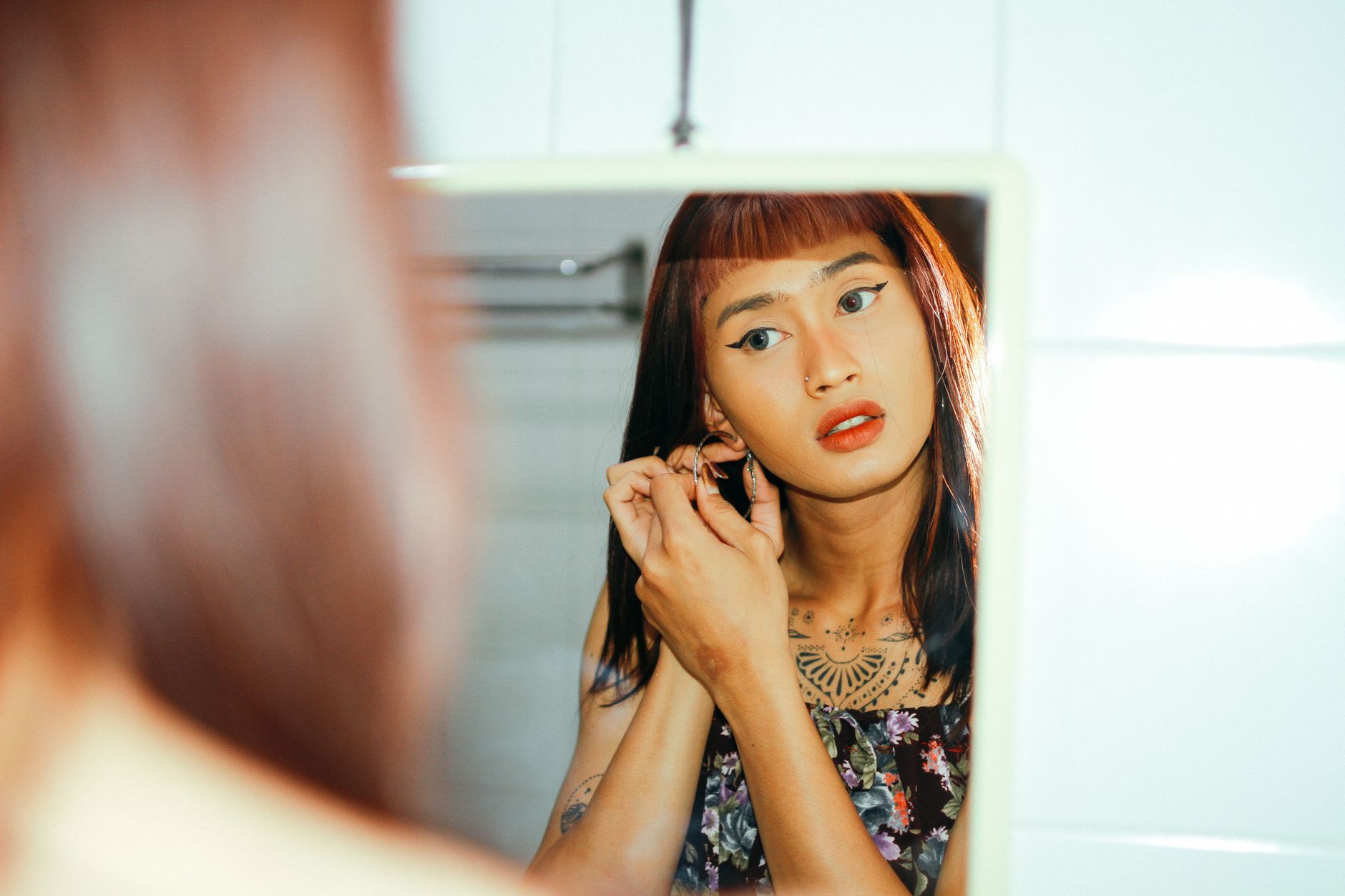 Asian woman with tattoos adjusting earring in front of a mirror indoors