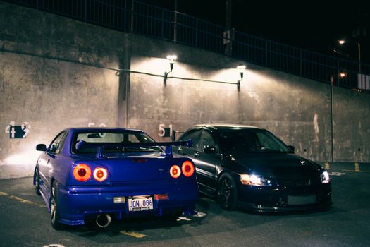 Two modified sports cars parked under streetlights in an urban parking lot at night.