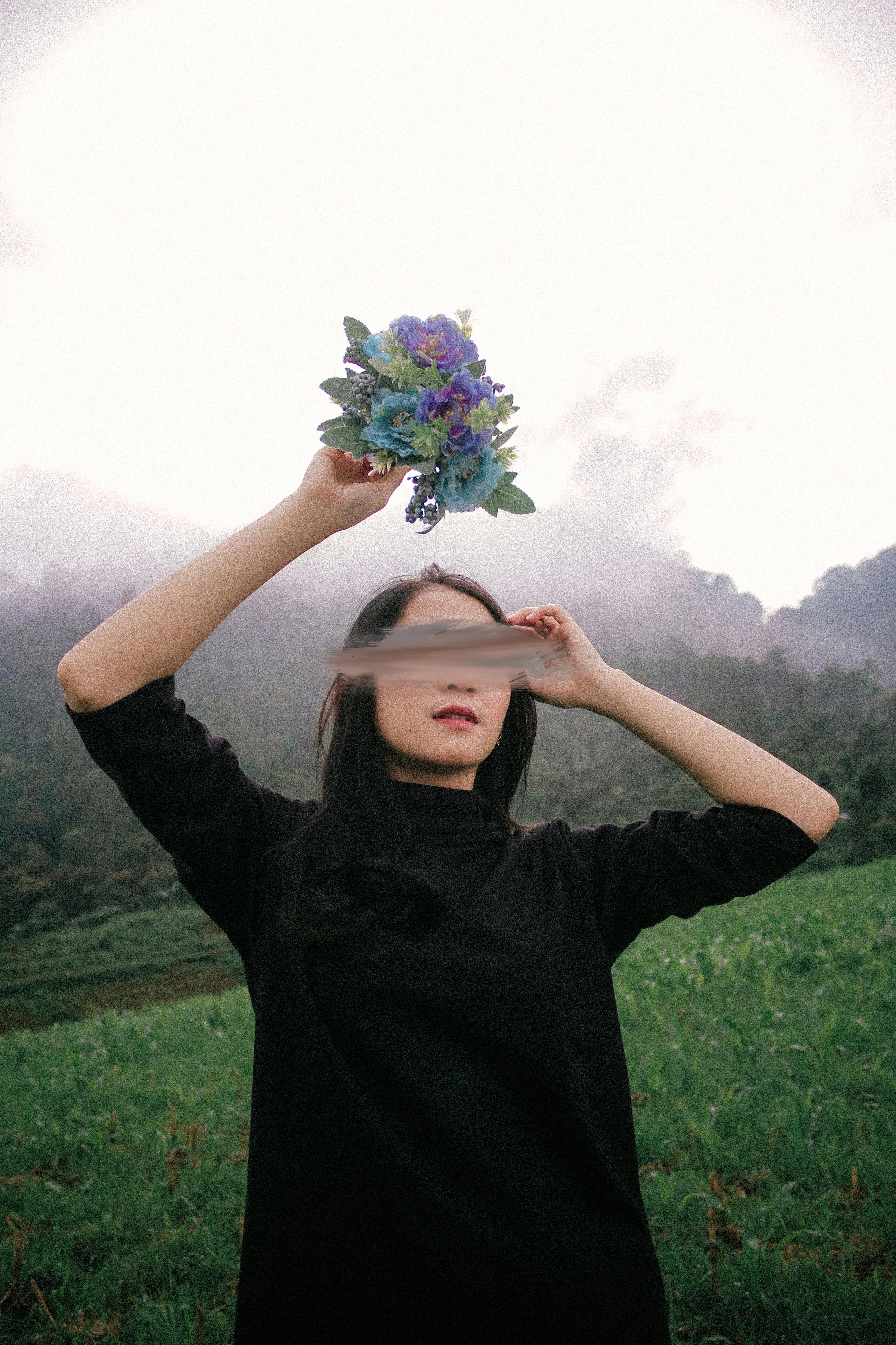 A woman in black raises flowers against a misty, green outdoor backdrop.