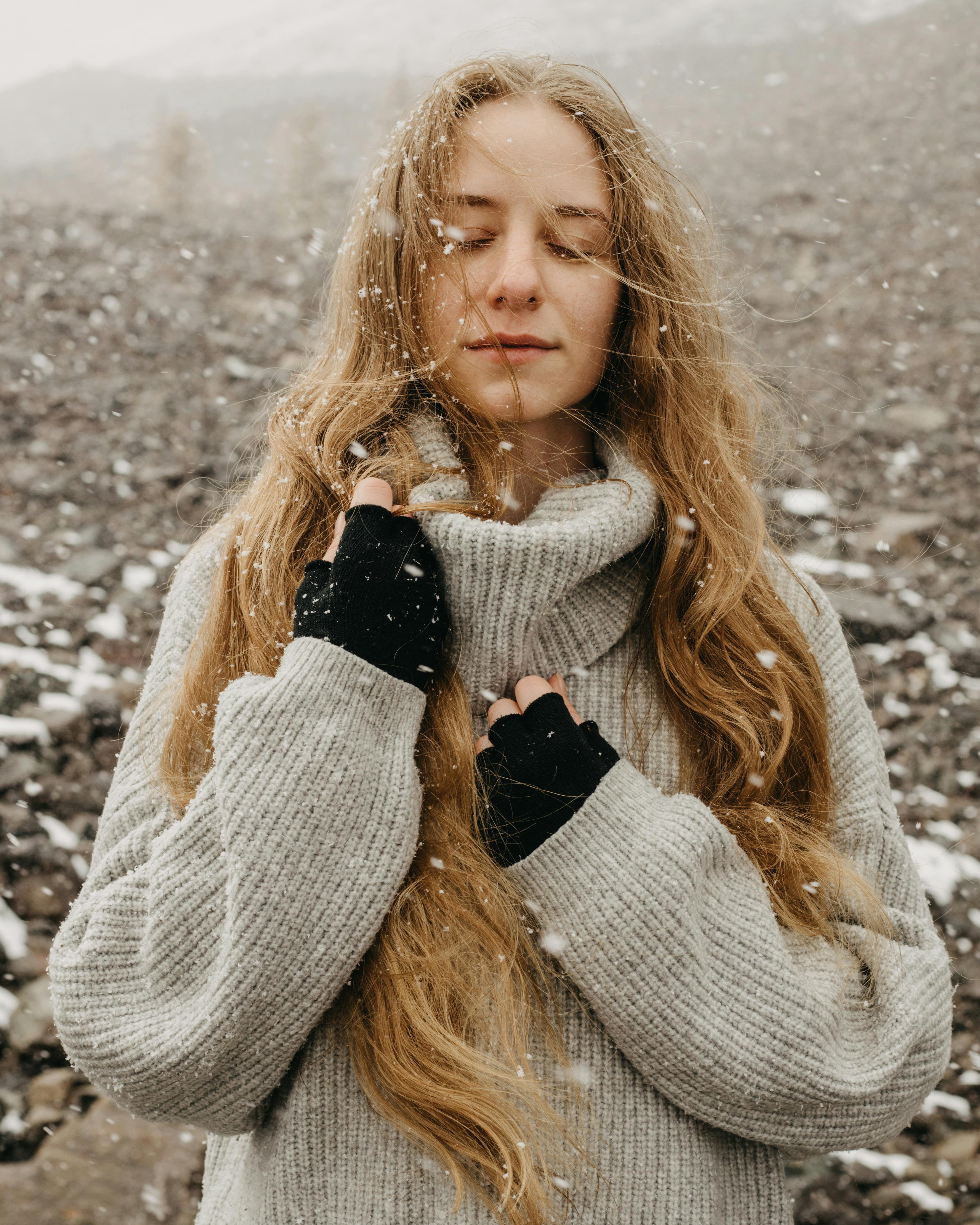 Serene woman with long hair in snowfall, wearing a cozy knit sweater and gloves.