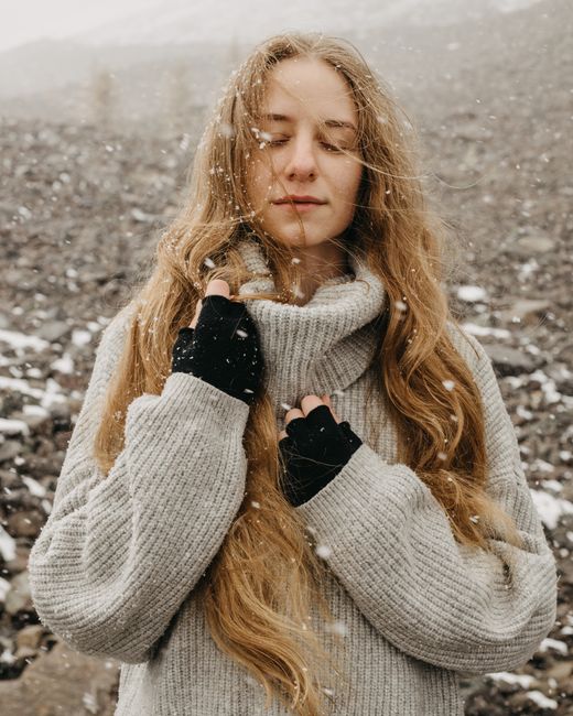 Serene woman with long hair in snowfall, wearing a cozy knit sweater and gloves.